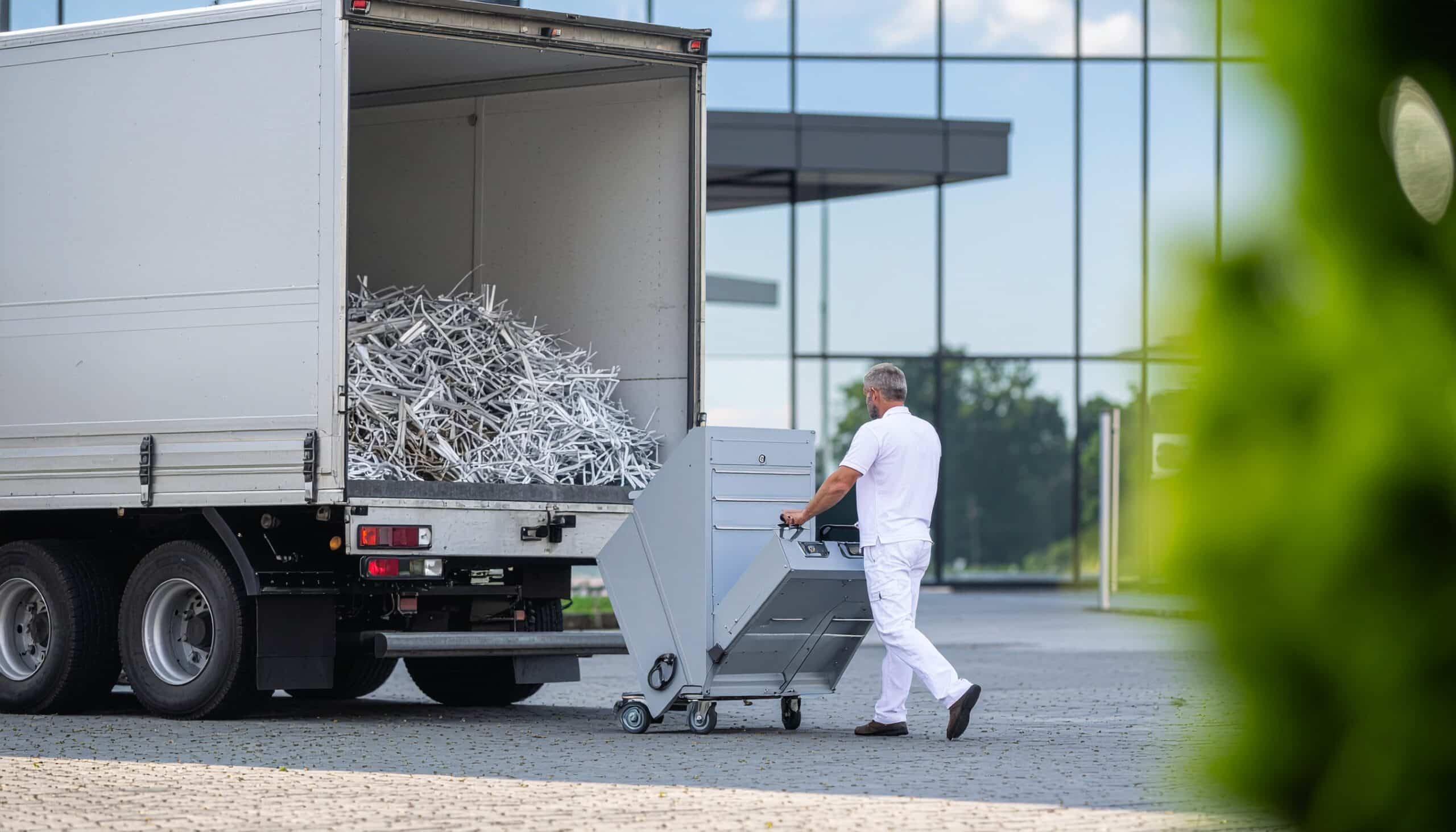 A person in white clothing is pushing a cart towards the back of a truck filled with metal scraps, with an industrial building in the background.